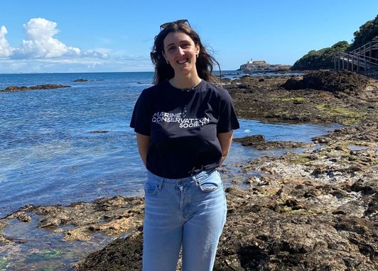 A member of Marine Conservation Society standing near some rockpools smiling at the camera. They are wearing a branding t-shirt and light blue jeans.