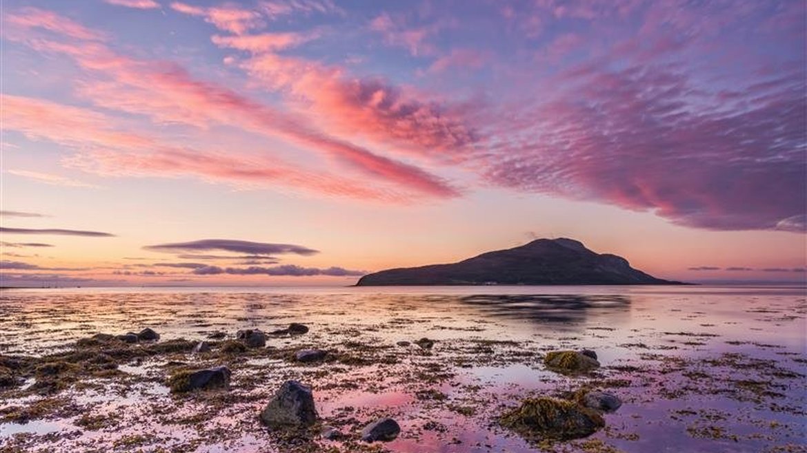 A view of Holy Isle from Lamlash Bay, Scotland. It is dusk and the sky is a beautiful mix of blue and purply pink clouds. The colours are reflected in shallow rock pools in the foreground.