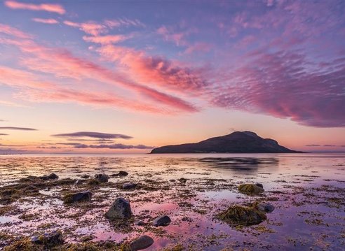 A view of Holy Isle from Lamlash Bay, Scotland. It is dusk and the sky is a beautiful mix of blue and purply pink clouds. The colours are reflected in shallow rock pools in the foreground.