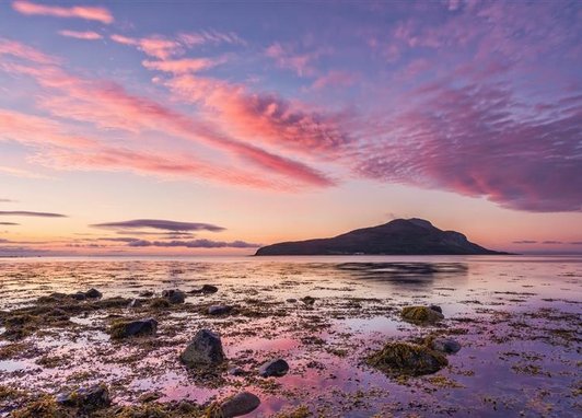A view of Holy Isle from Lamlash Bay, Scotland. It is dusk and the sky is a beautiful mix of blue and purply pink clouds. The colours are reflected in shallow rock pools in the foreground.