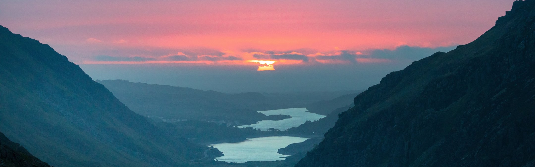 The sun is pictured rising from the top of Mount Snowdon. The sky is pink and orange as the sun is slowly rising, its beams reflected on the water in the valley