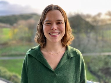 Trustee polly Burns smiles against a green landcaspe. She has short wavy brown hair and is wearing a moss-green jumper.