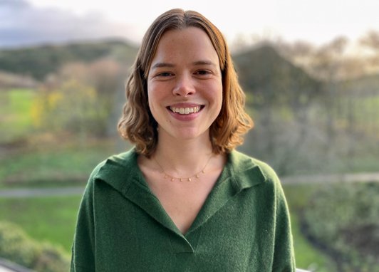 Trustee polly Burns smiles against a green landcaspe. She has short wavy brown hair and is wearing a moss-green jumper.