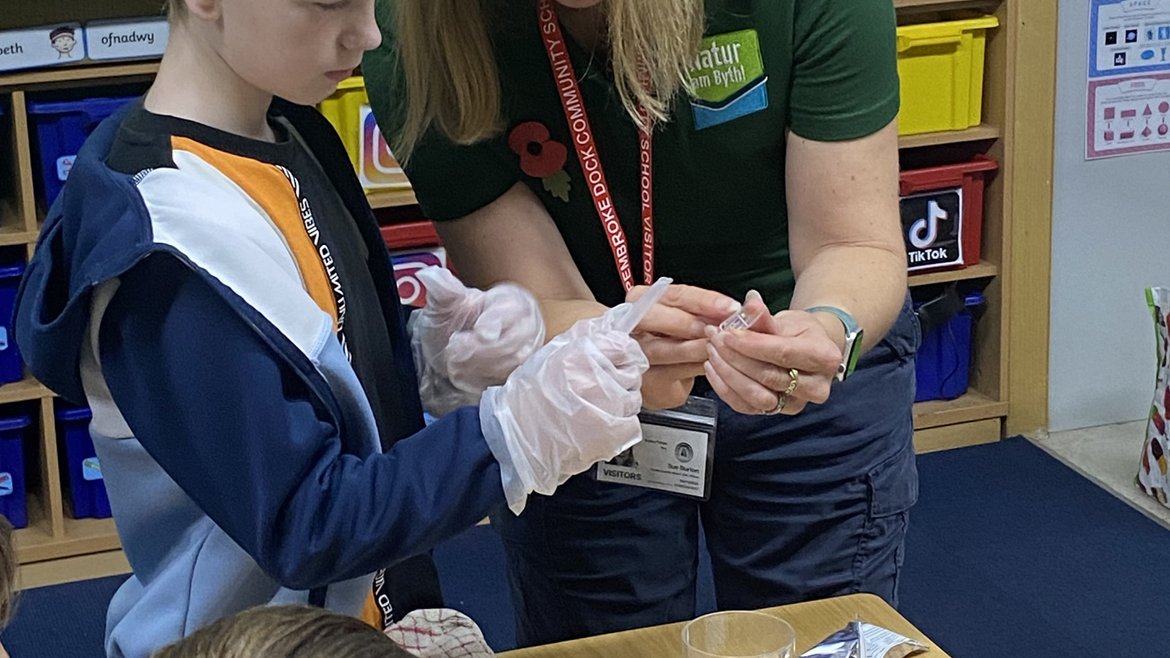 Sue Burton stands next to a schoolchild in a classroom. She is showing them a vial of water. On the table in front of them are beakers of water and water quality checking equipment