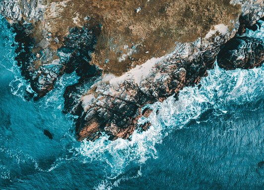 An aerial view of a rugged, rocky coastline where turquoise waves are crashing against the shore.