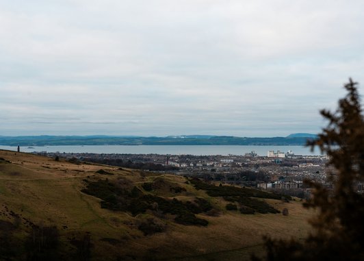 A wide-angle, slightly elevated view overlooking the city of Edinburgh, Scotland, under a grey, overcast sky. In the foreground, rolling green and brown hills slope downward.