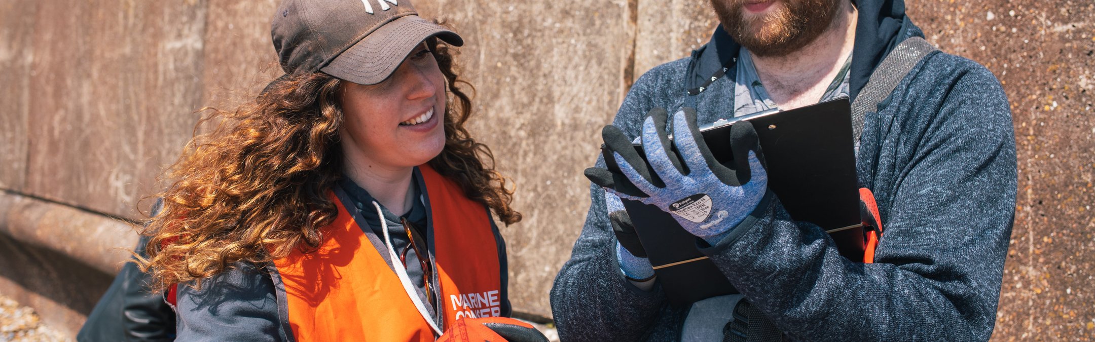 A woman with long, curly brown hair wearing a brown cap, orange high-vis vest and orange work gloves points toward a clipboard held by a man. The man is wearing a blue hoodie and blue work gloves. They are standing outdoors in front of a concrete wall.
