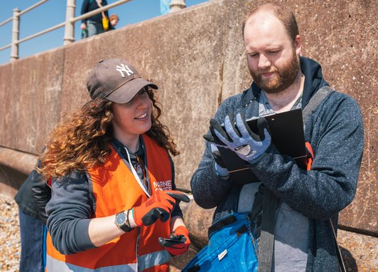 A woman with long, curly brown hair wearing a brown cap, orange high-vis vest and orange work gloves points toward a clipboard held by a man. The man is wearing a blue hoodie and blue work gloves. They are standing outdoors in front of a concrete wall.