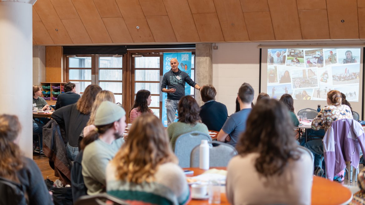 A member of the Ocean Learning team speaking to a group of residents. On the whiteboard is a presentation on volunteering.