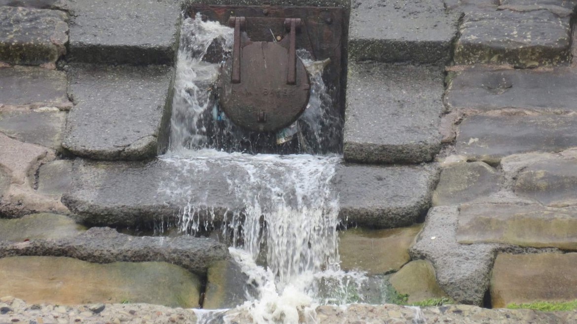 Dirty water is flowing from a rusty metal flap gate in a concrete and stone wall, it is splashing onto the beach below