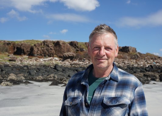 Trustee Mark Eckstein smiles on a sandy beach with a rocky bank in the background and a blue, slightly cloudy sky. He is wearing a checked shirt.