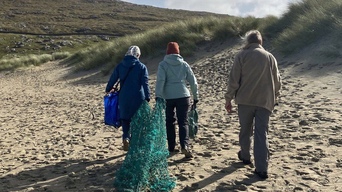 Three volunteers walk away from the camera along the sand at Luskentyre beach holding a large green fishing net behind them