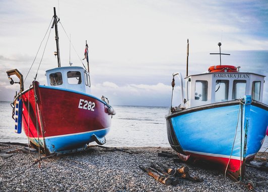 A close up of two fishing boats resting on pebbles. One boat is a vivid red colour, and the other is a bright blue colour.