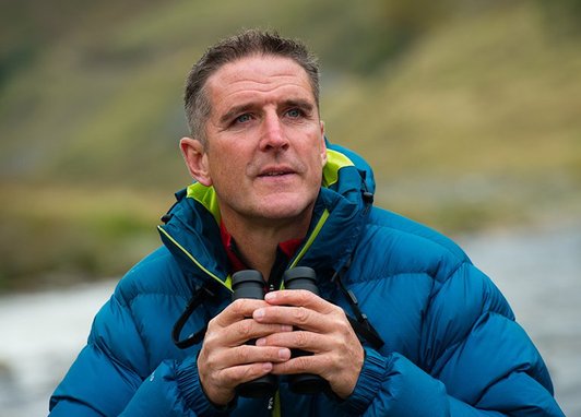 Iolo Williams stands against a Welsh mountain backdrop wearing a blue puffer coat. He is holding binoculars.