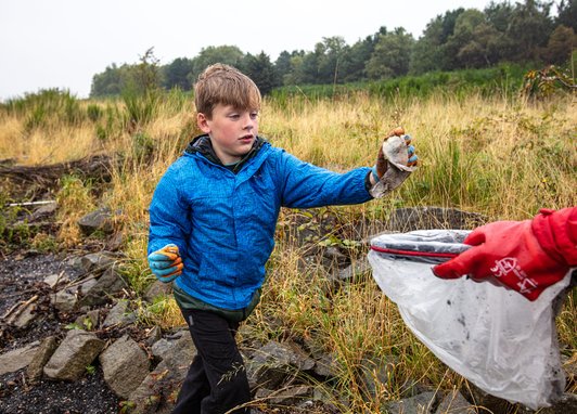 A young boy in a blue jacket and red gloves using a litter picker to collect rubbish. He is putting a soiled wet wipe into a white bag which is being held by someone off camera.