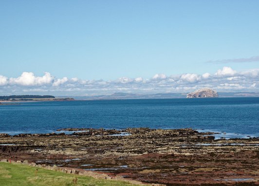 A view out over the Firth of Forth with a rocky shore in the foreground and a beautiful view across the water. The sky is blue and the clouds sit low in the sky.