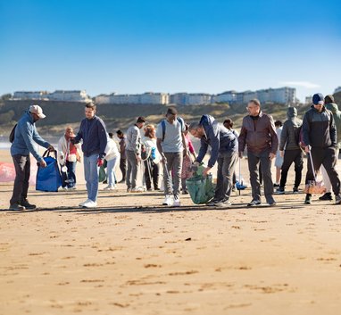 A group of beach cleaners holding waste bags and litter pickers on a snady beach. The sun is shining off camera, leaving shadows on the beach under them.