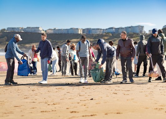 A group of beach cleaners holding waste bags and litter pickers on a snady beach. The sun is shining off camera, leaving shadows on the beach under them.