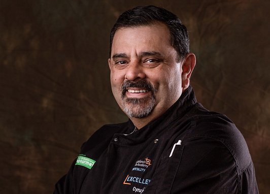 Chef Cyrus Todiwala smiles at the camera wearing chef's blacks against a black background.