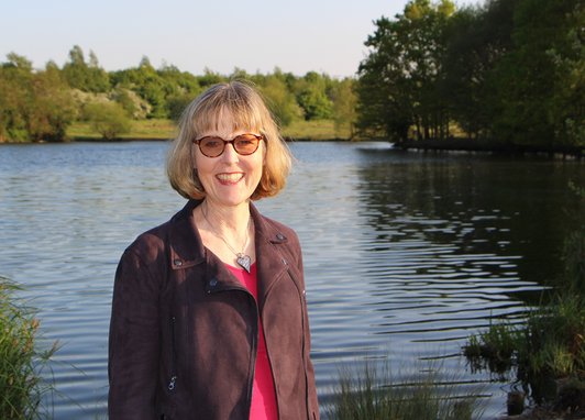 A picture of a smiling Amanda Nobbs, the Marine Conservation Society's Chair. She is standing in front of a calm lake backed with trees and has short blonde hair, a brown jacket and pink shirt.