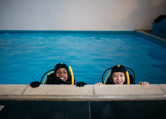 2 children learning to dive at Dive Project Cornwall. They are in a pool wearing diving gear and smiling at the camera