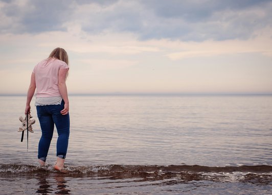a person standing at the edge of the sea while holding a pair of shoes