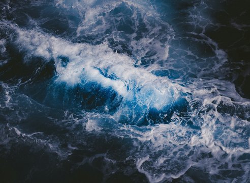An aerial view of dark ocean waves. The water is indigo blue and white peaks of crashing waves can be seen.
