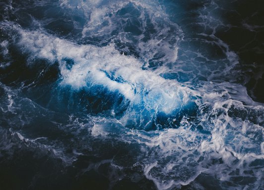 An aerial view of dark ocean waves. The water is indigo blue and white peaks of crashing waves can be seen.