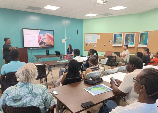 A room of adults listening to a presentation being given. The screen reads "Fishing"