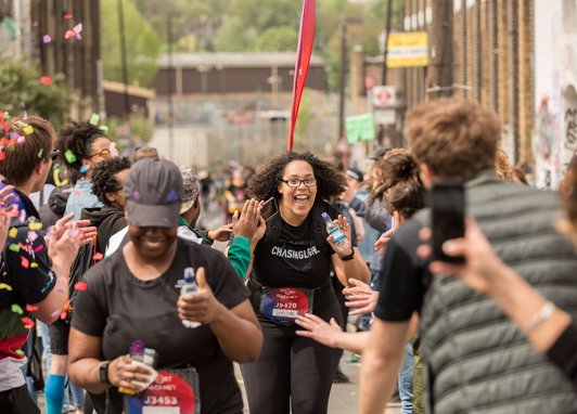 A happy runner with curly hair and glasses, wearing a black t-shirt, celebrates as she approaches a finish line. She is surrounded by supporters