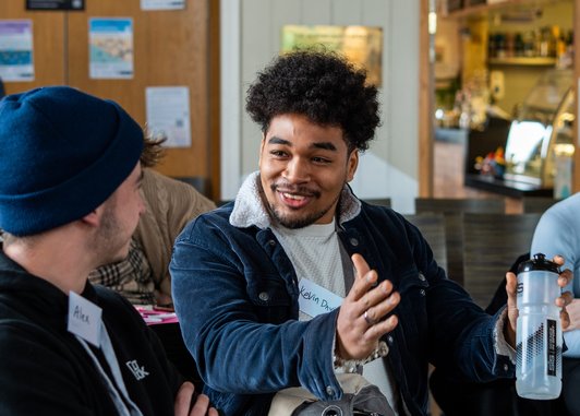 A young man with dark curly hair is wearing a blue corduroy jacket with a white fleece collar, he is smiling and gesturing with his hands while talking to another person. The person he's speaking to is wearing a blue beanie and a black hoodie.