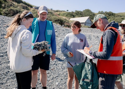 Marine Conservation Society staff working with young people on a beach. One girl is holding a large litter item, while a staff member fills out the litter survey paper form.