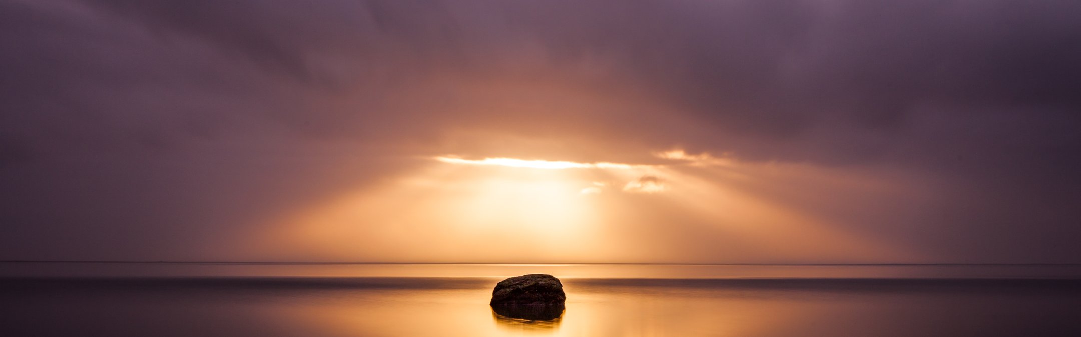 A sunset over a coast. The sun is hidden behind dark, grey clouds. Rays are hitting the water and causing a solitary rock on the water to be silhouetted.