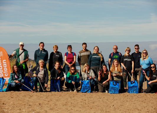 A group of 18 volunteers, some standing and some crouching, pose on a sandy beach with blue collection bags and litter pickers after a beach clean event as part of the Marine Conservation Society's Beachwatch program