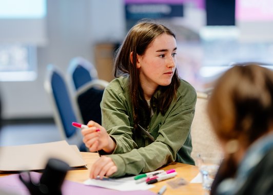 A young adult sitting at a table looking to their left away from the camera. They are holding a pen in their right hand.