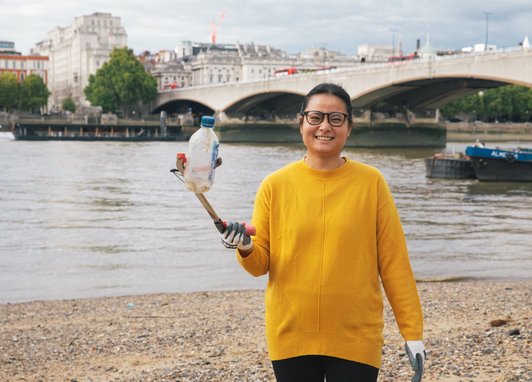 A woman in a yellow jumper holding up a plastic bottle to the camera using a litter picker. She is in front of the river at Festival Pier, London