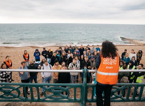 A woman with her back to the camera speaking to a group of beach cleaners all huddled in front of her on the beach. The woman is wearing a Marine Conservation Society vest.