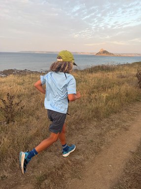 A young boy running along a dirt path. The coast is behind him. He has his back away from the camera and is weiring a lime green cap and blue t-shirt.