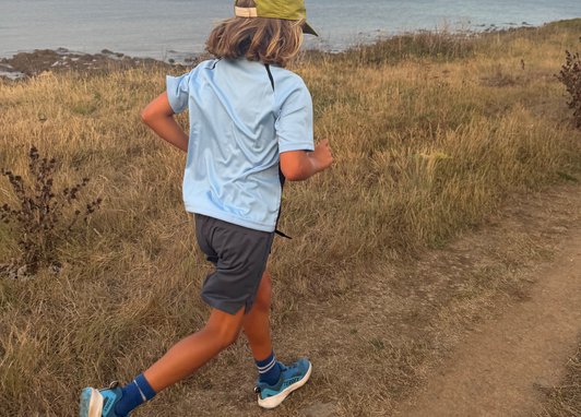 A young boy running along a dirt path. The coast is behind him. He has his back away from the camera and is weiring a lime green cap and blue t-shirt.