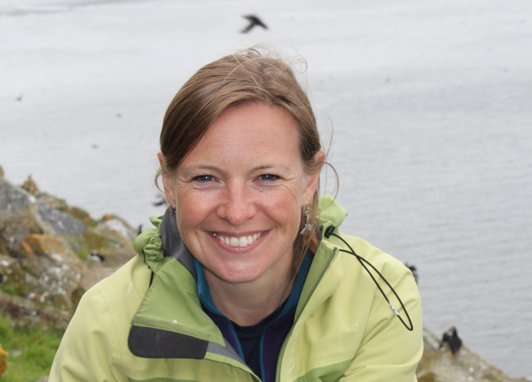 Miranda Krestovnikoff smiles at the camera from a rocky cliff face with the sea in the background. It appears there are puffins flying in the background, although their silhouettes are small.