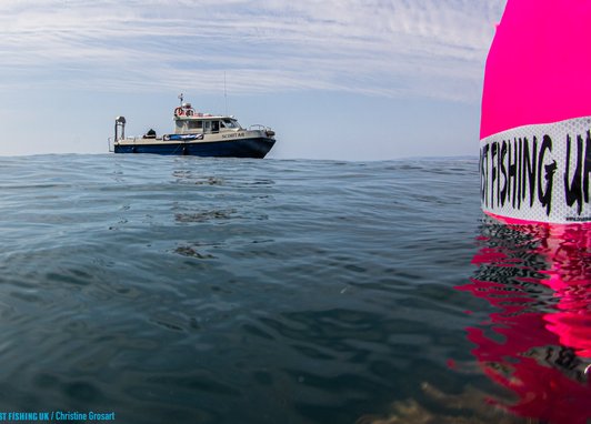 A fishing boat in shown in the distance with a bright pink buoy-type device reading 'ghost fishing UK' in the foreground.